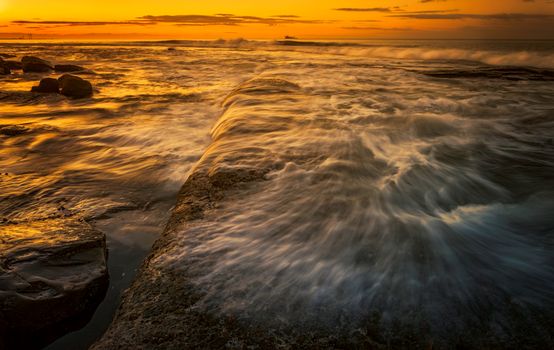 Dramatic long exposure of ocean waves crashing against rocks at sunset.