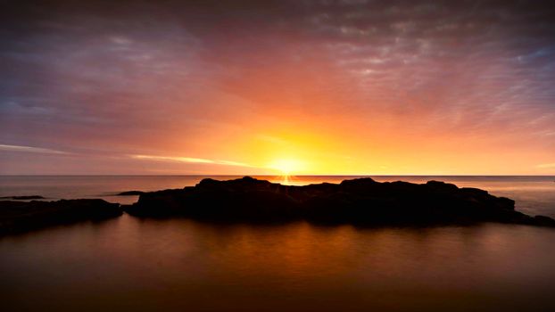 Stunning ocean sunset with dramatic sky and rock formations in silhouette.