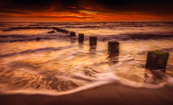 Dramatic ocean waves crash against wooden posts during a vibrant sunset.