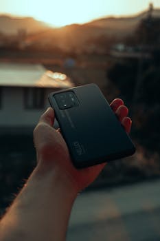 Close-up of a hand holding a smartphone outdoors with a serene sunset in the background.