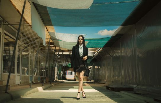 Confident woman in stylish attire walking through a sunlit outdoor corridor.