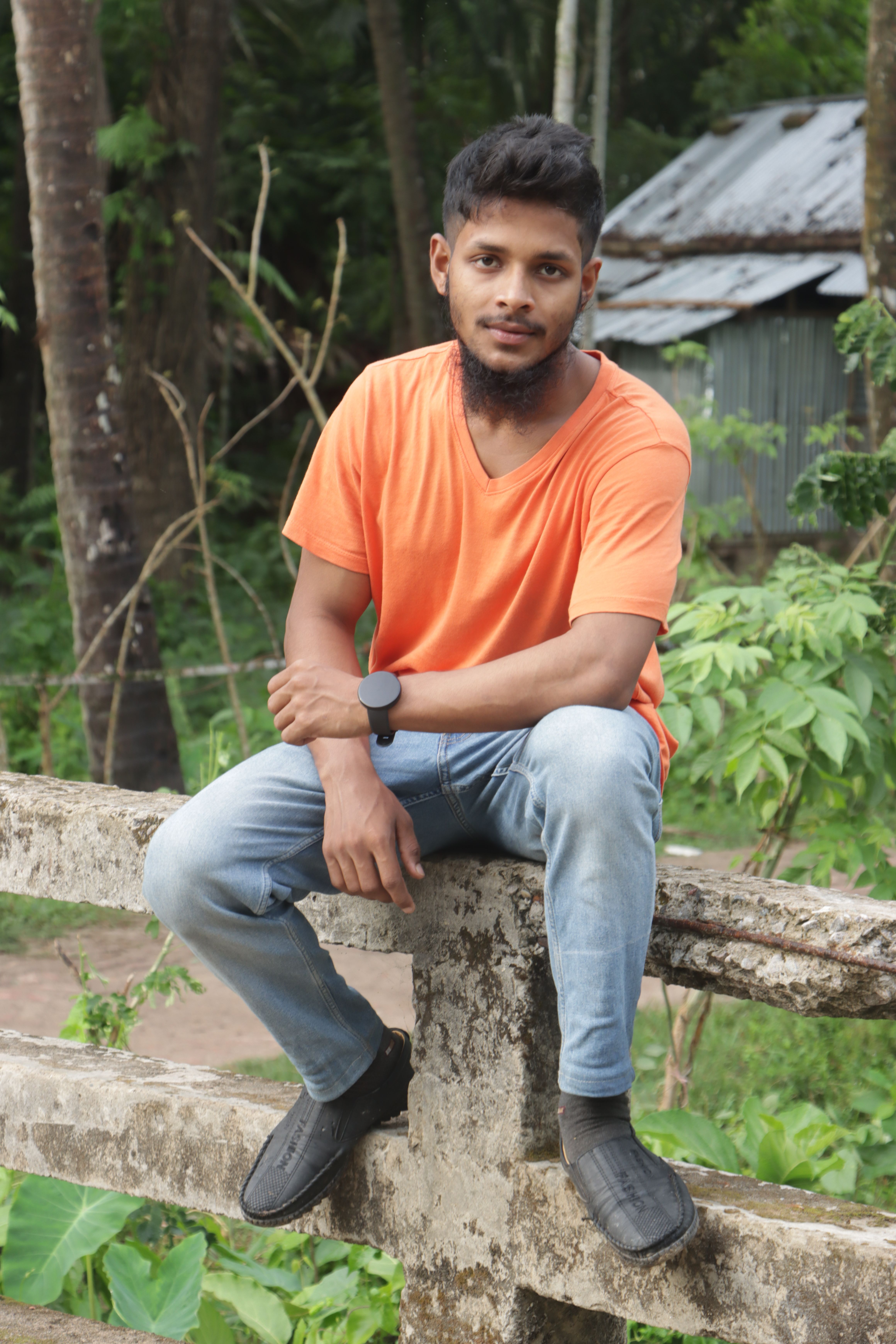 A young man in an orange shirt sits on a fence in a lush outdoor setting.