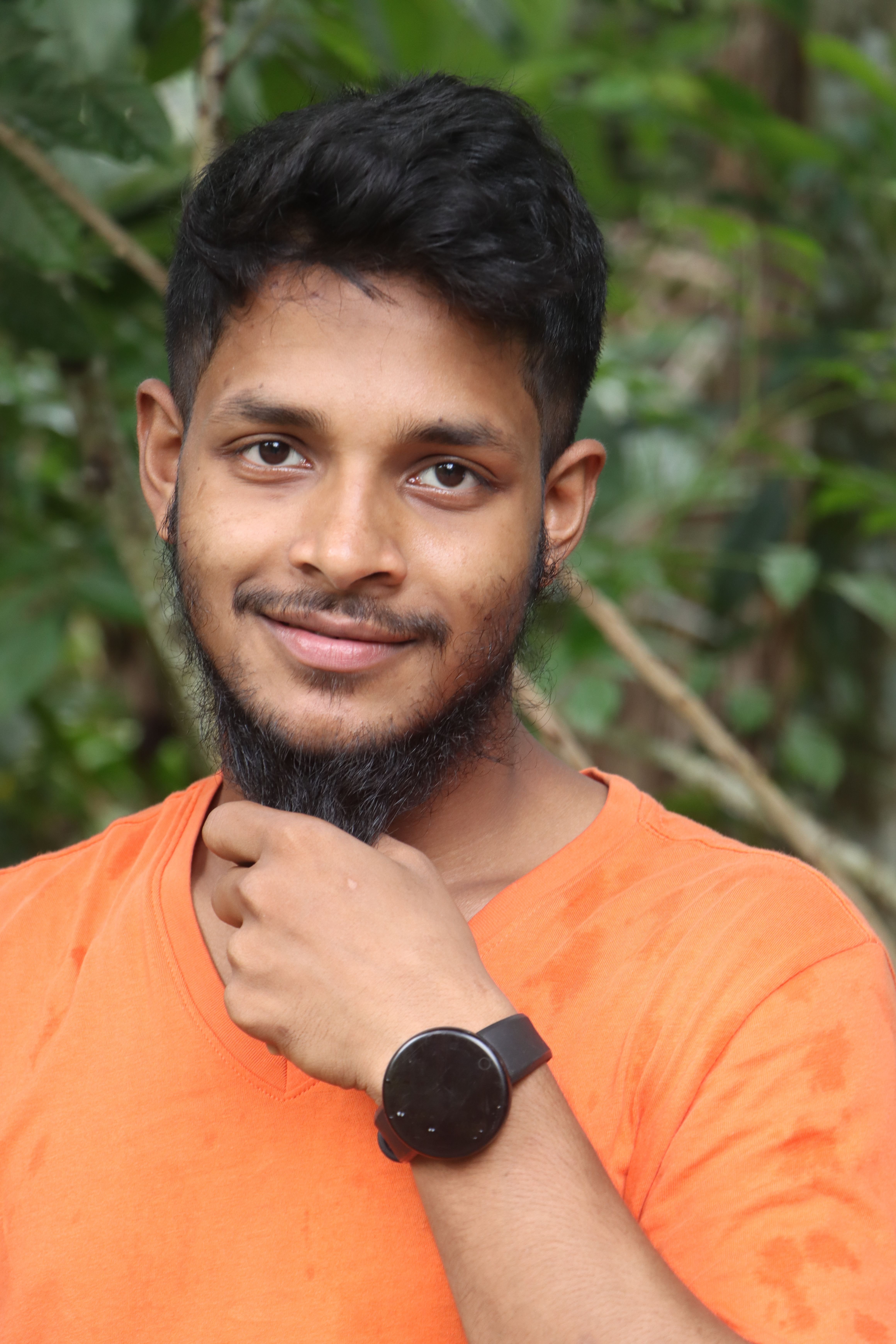 A bearded young man smiles warmly while wearing an orange shirt and watch in a lush green outdoor setting.