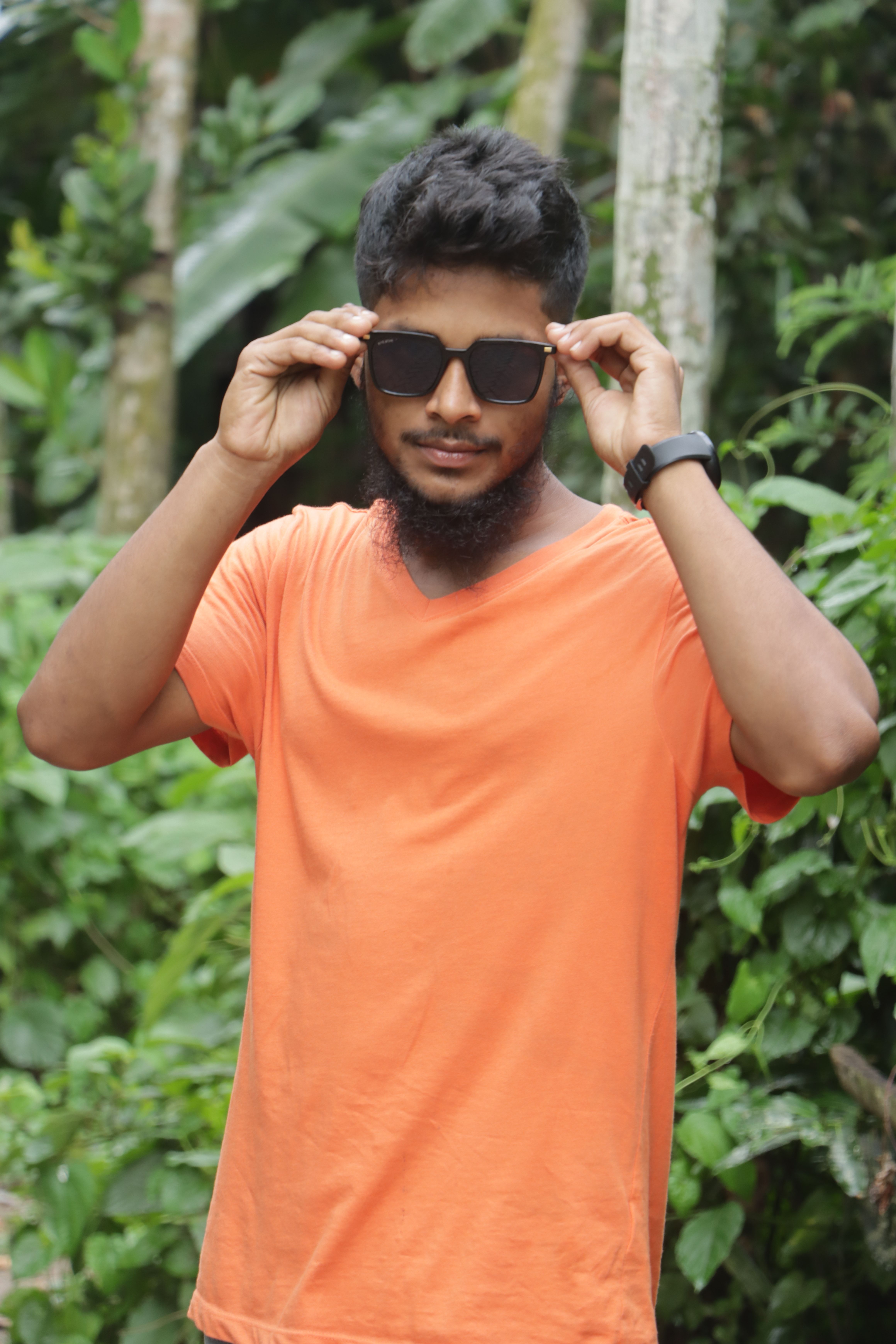 Young stylish man outdoors in an orange shirt adjusting sunglasses amidst lush greenery.