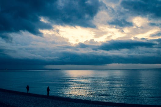 Silhouetted figures on a beach under a dramatic cloudy sky at dusk, serene ocean view.