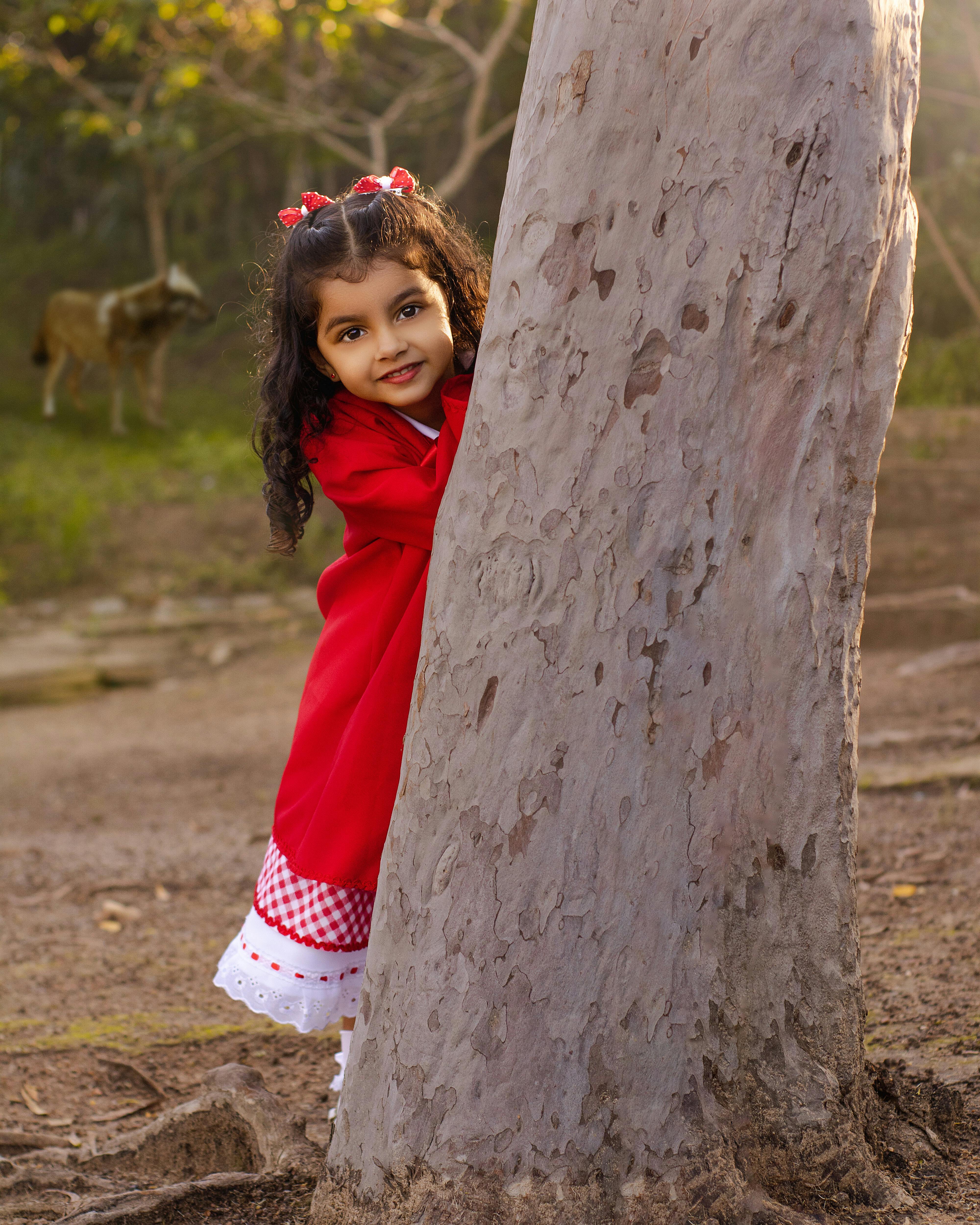 Cute girl in red dress playing behind a tree in a sunny park
