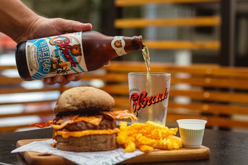 A Person Pouring Beer into a Glass Next to the Burger and Fries 