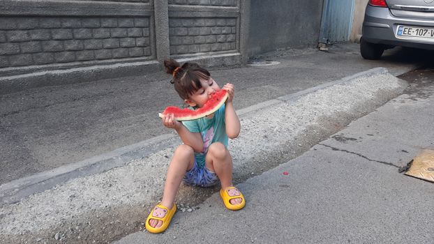 A young girl sits on a sidewalk eating a juicy watermelon slice on a city street.