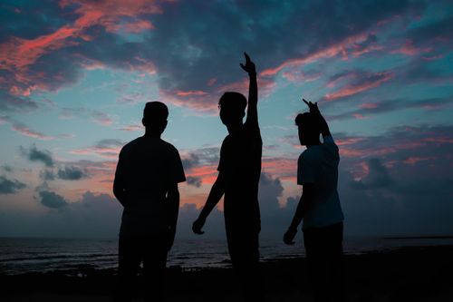 Free Three friends enjoy a colorful sunset, silhouetted against the sea with hands raised in celebration. Stock Photo