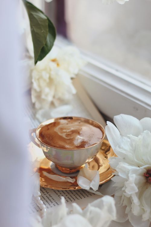 Free A steaming cup of coffee on a golden saucer surrounded by white flowers by a windowsill. Stock Photo
