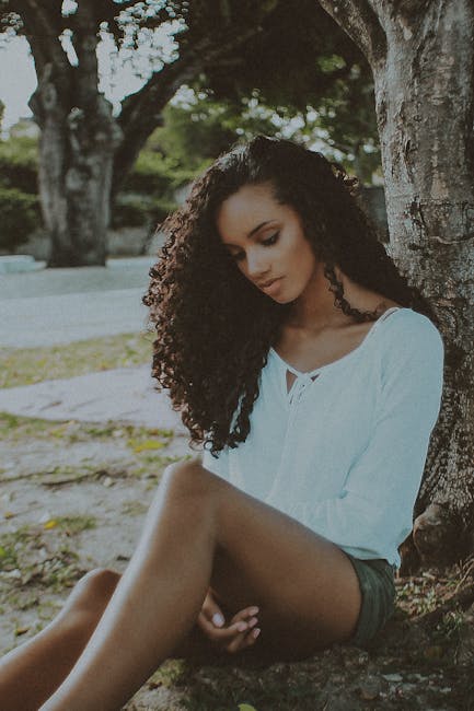 A contemplative woman with curly hair sits outdoors under a tree in a serene park setting.
