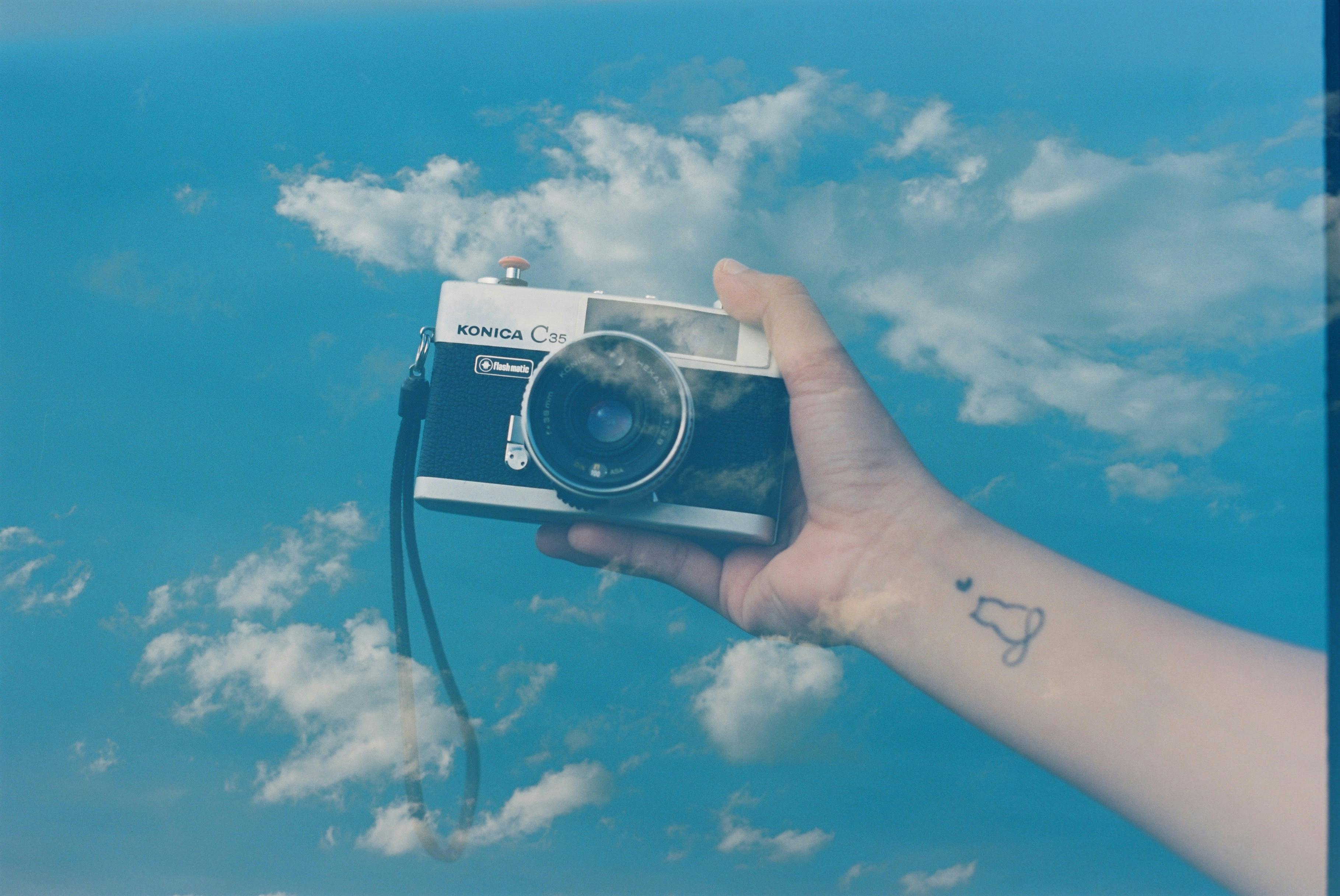 Artistic double exposure of a vintage camera held by a hand against a blue sky with clouds.