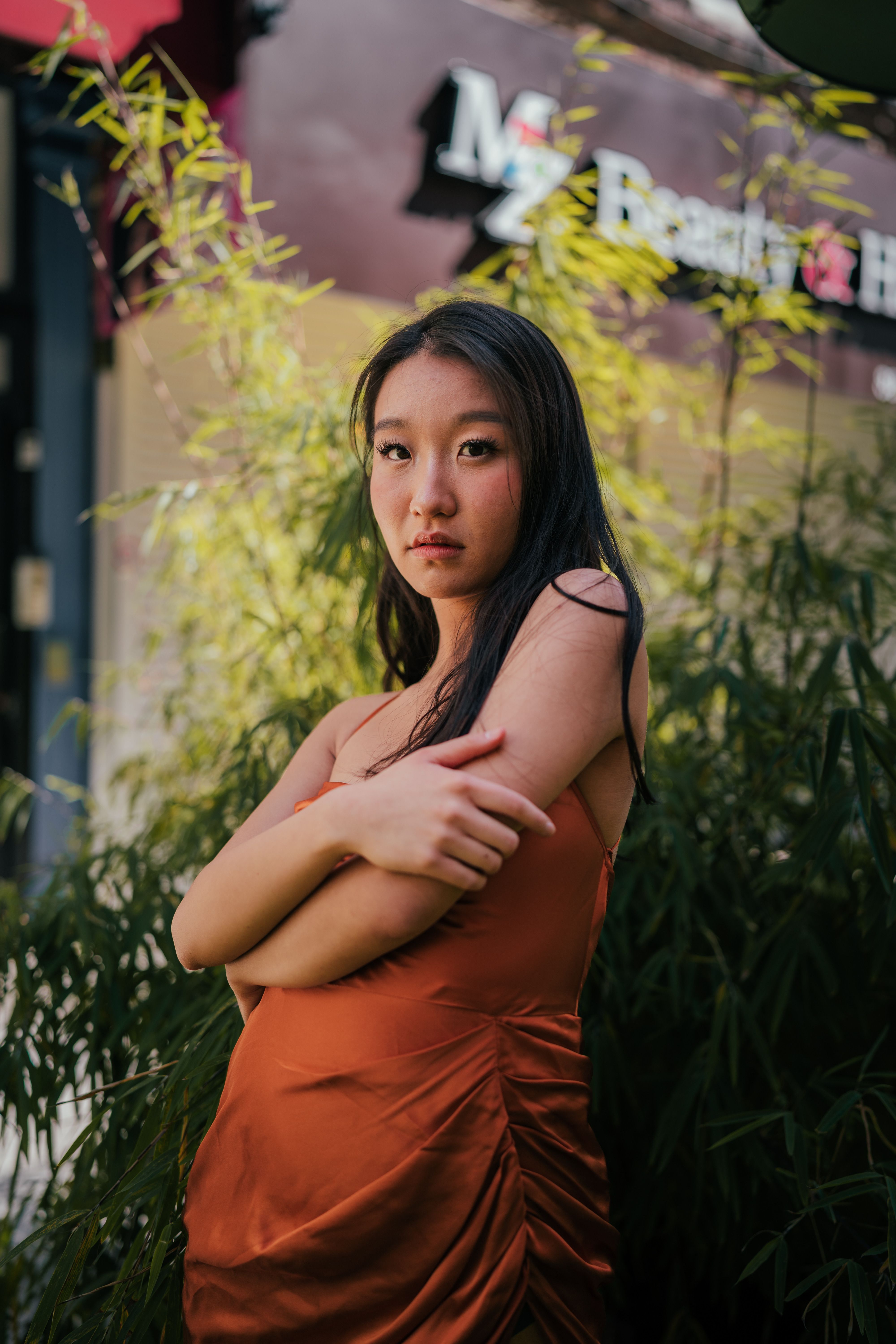 Young woman in orange dress posing elegantly among green foliage outdoors.