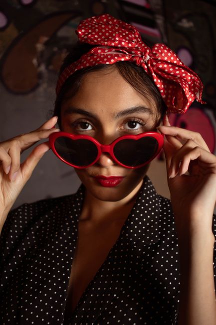 Fotomural Mujer elegante con gafas de sol en forma de corazón y vestido de lunares, evocando un ambiente vintage en un entorno moderno.