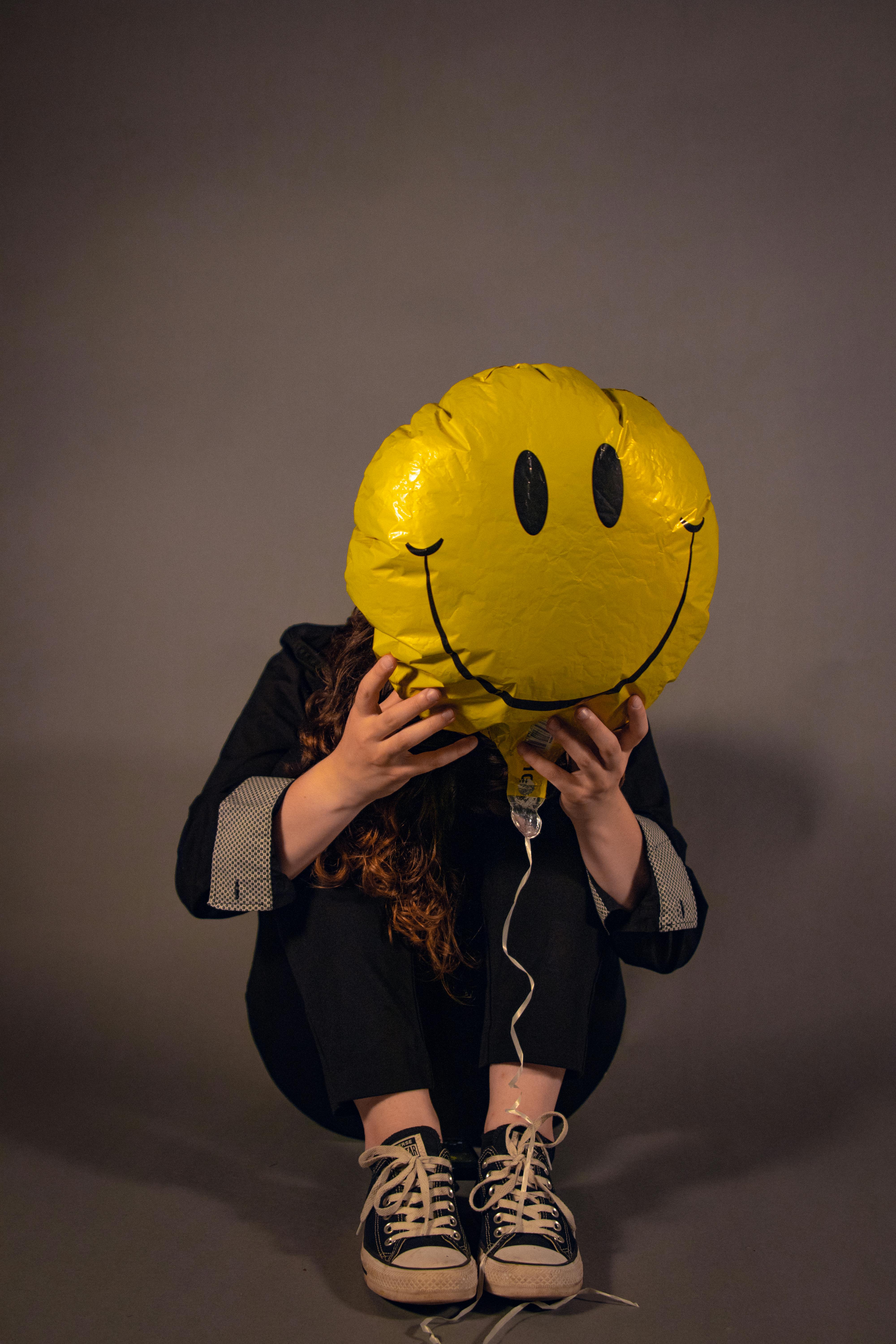Teenager sitting in studio holding a large smiley face balloon, creating an artistic and playful vibe.