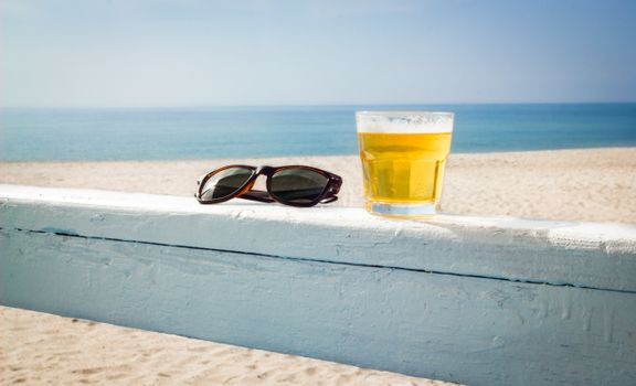 A chilled glass of beer and sunglasses on a beach railing. Perfect summer vibes.
