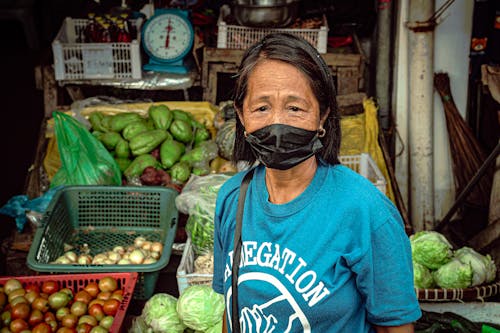 A Portrait of an Elderly Woman Wearing a Blue Shirt and a Face Mask at a Market