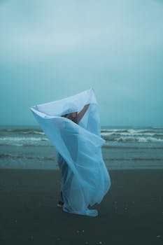 A woman stands with flowing fabric on a cloudy beach by the ocean, creating a serene scene.
