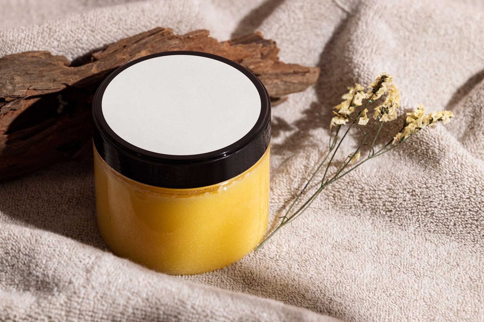 Jar of yellow skincare cream on a towel with wood and dried flowers, ideal for spa themes.