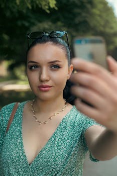A young woman takes a selfie with her smartphone in a sunny outdoor park setting during summer.