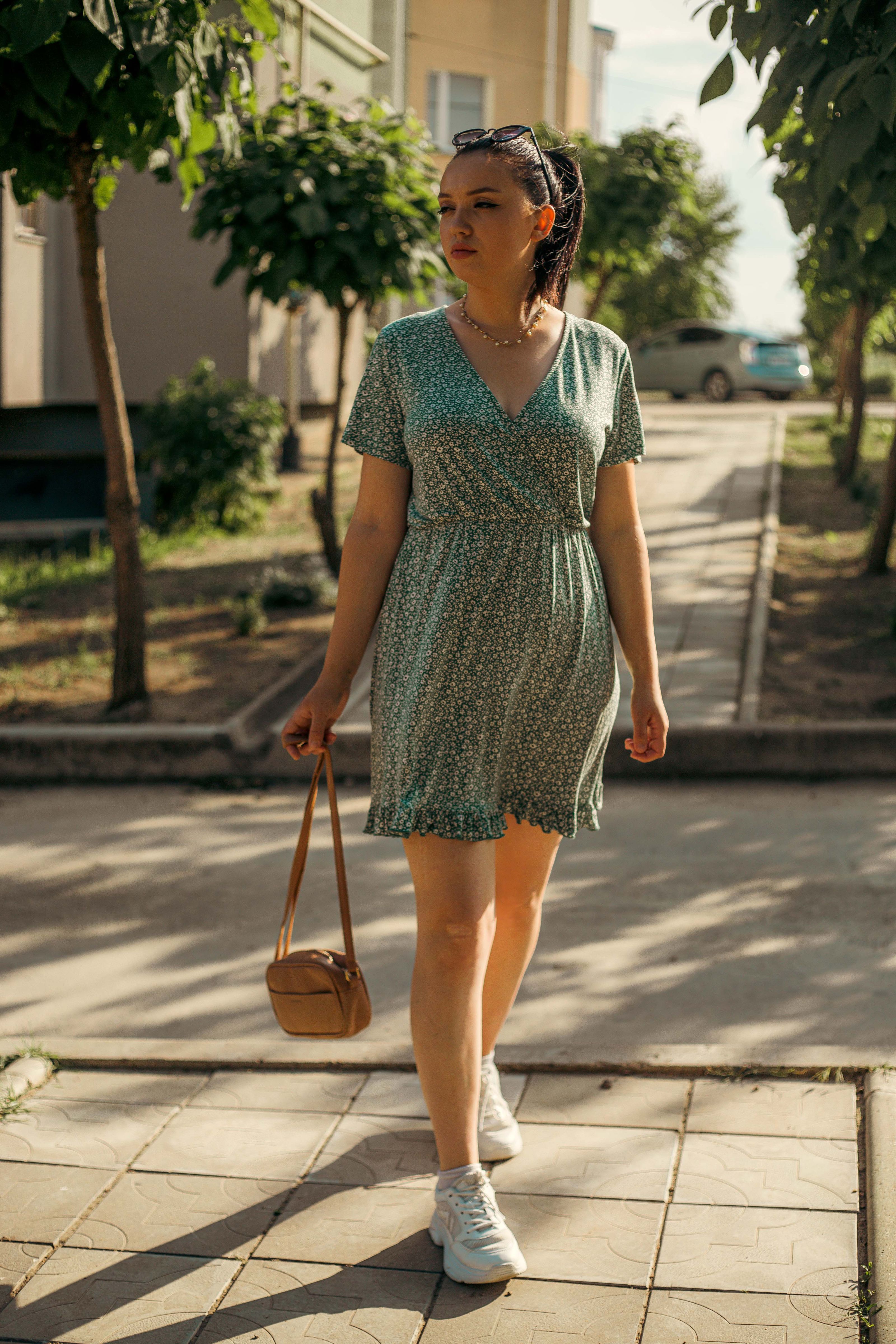 Uma jovem em um vestido verde anda em uma calçada ensolarada, carregando uma bolsa marrom.