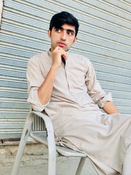 Young man in traditional attire poses thoughtfully indoors against a corrugated metal wall.