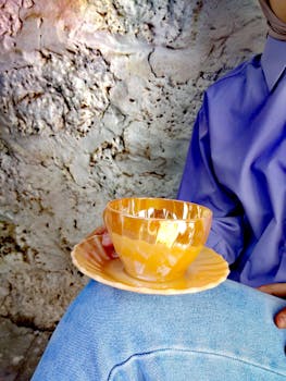 A person in blue holds a shiny golden cup and saucer against a stone wall.