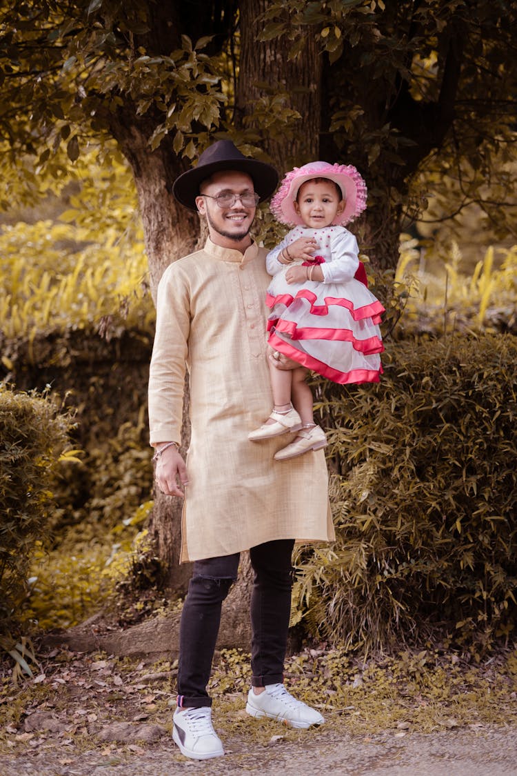 Man In Beige Kurta With Black Hat Carrying Baby Girl In Pink And White Dress