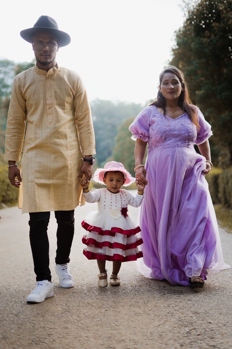 Man In Beige Kurta And Woman In Purple Dress Walking Hand In Hand With Girl In Pink And White Dress