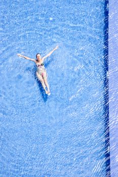 Aerial shot of a woman swimming and relaxing in a bright blue swimming pool on a sunny day.