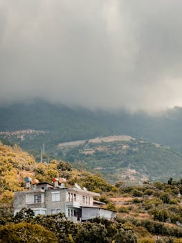 A tranquil mountain scene in Alanya, Turkey, with a house amid lush greenery under a foggy sky.