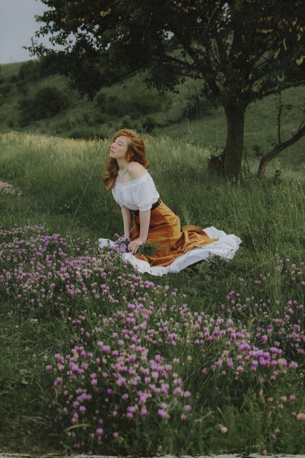 A woman in a vintage-style dress kneels amidst a field of pink flowers, embracing nature's beauty.