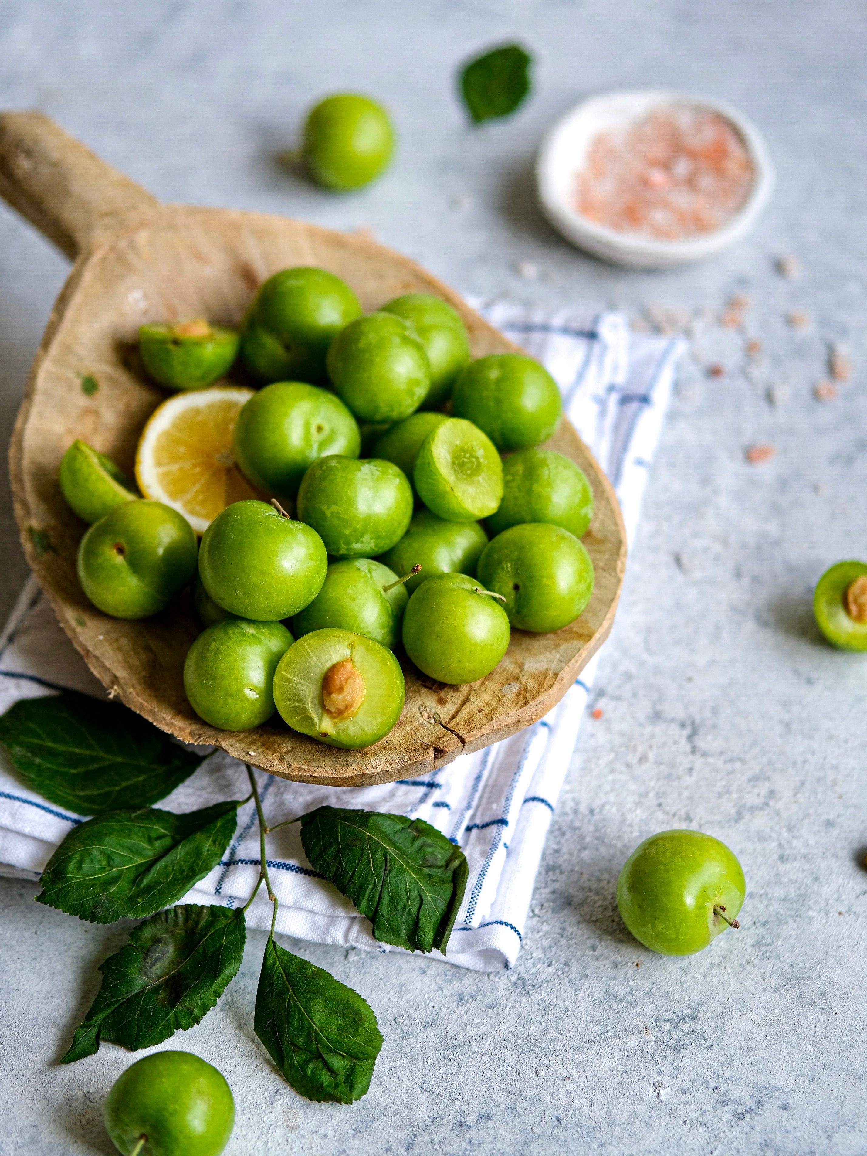Close-up of fresh green plums, lemon, and salt on a table with leaves, depicting a traditional Turkish snack.