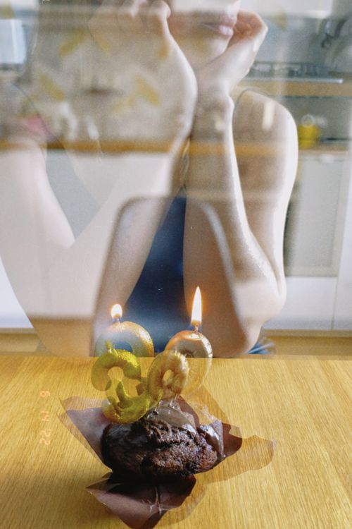 Free A woman reflected in glass sits contemplatively with a birthday cupcake and candles on a wooden table. Stock Photo