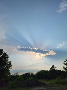 A breathtaking view of sunrays piercing through clouds, illuminating the serene blue sky above green trees.
