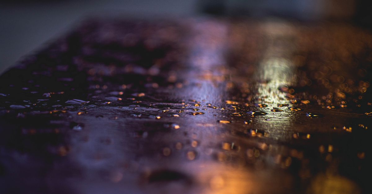 Imagem escura e sombria de gotas de chuva criando reflexos em uma superfície molhada.
