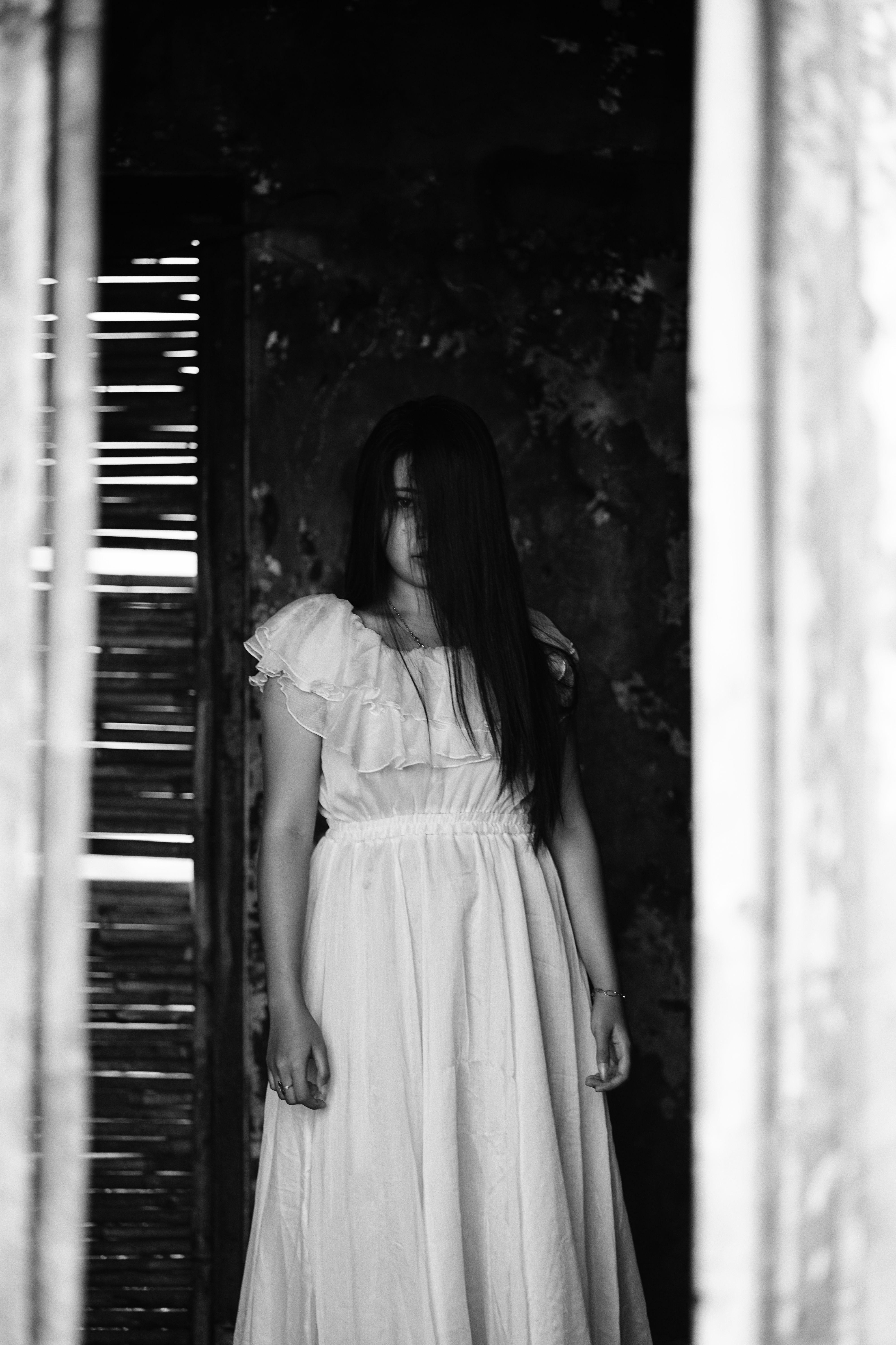 Black and white portrait of a woman in a flowing dress with long hair obscuring her face, in a dark, rustic setting.