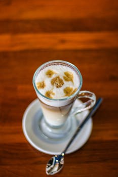 Close-up of latte macchiato with foam art in a transparent glass, placed on a saucer with a spoon, on a wooden table.