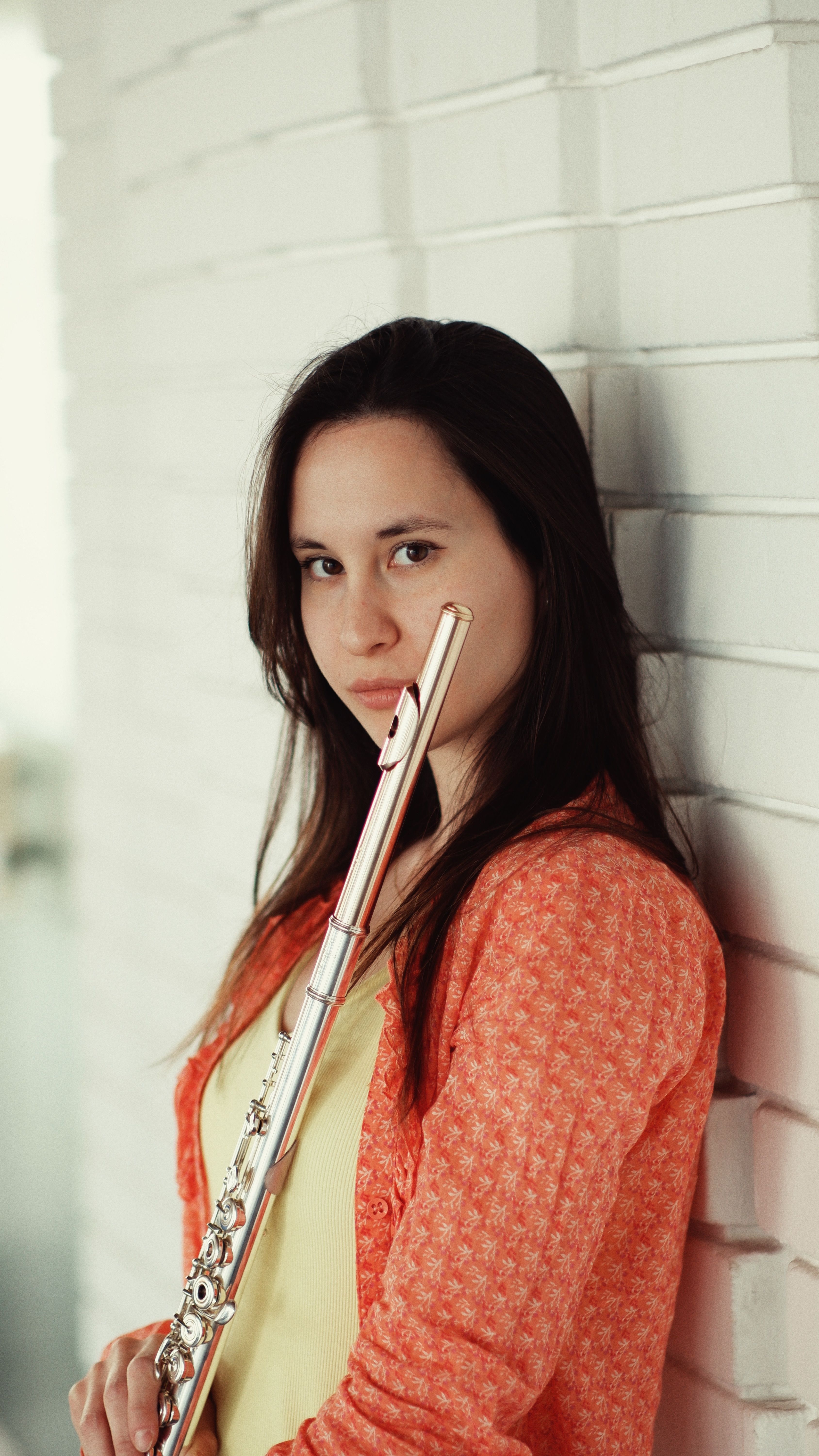 A woman leans against a white brick wall, holding a flute with a calm expression.