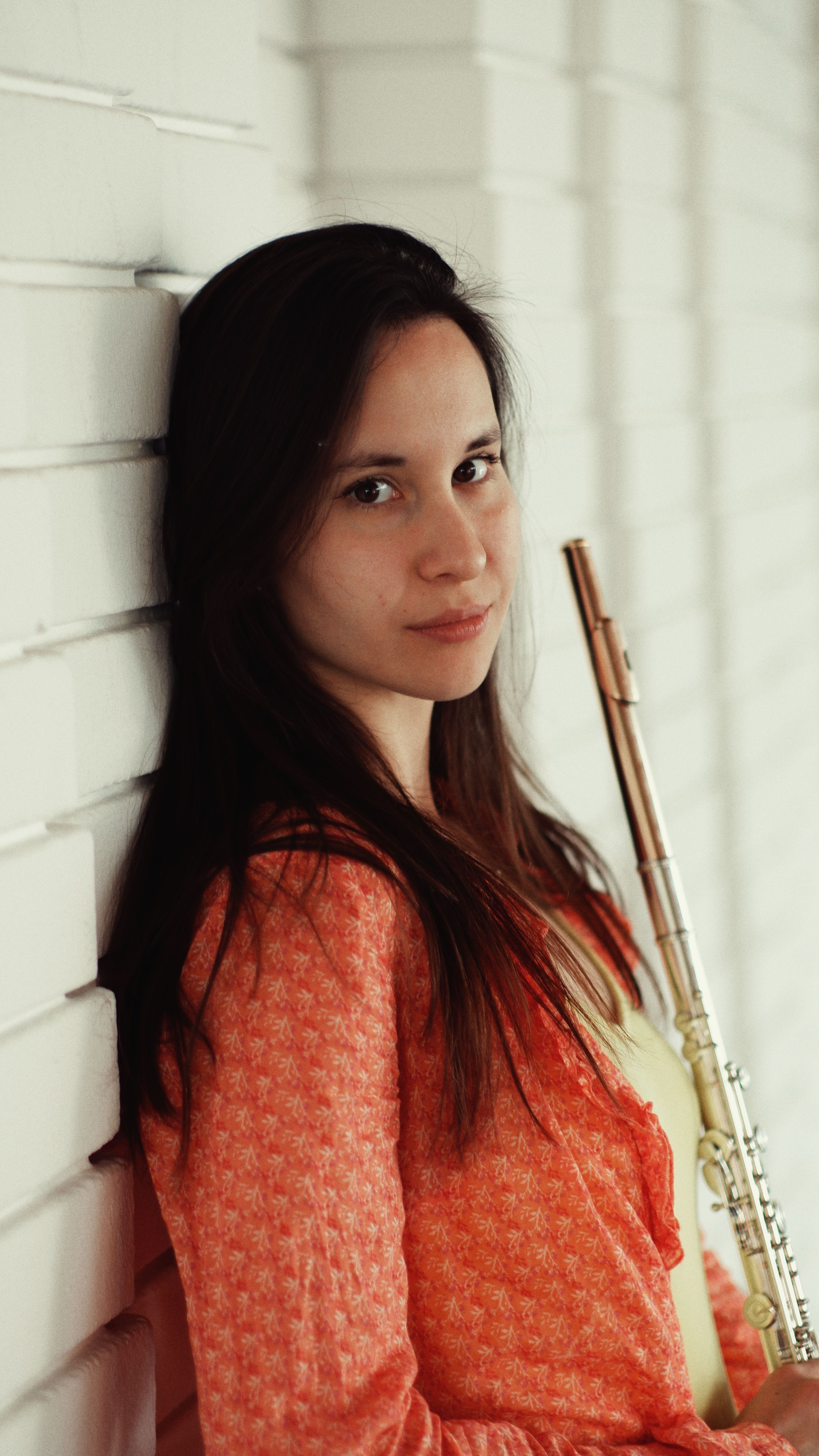 Portrait of a young woman holding a flute against a white brick wall, exuding elegance and passion.