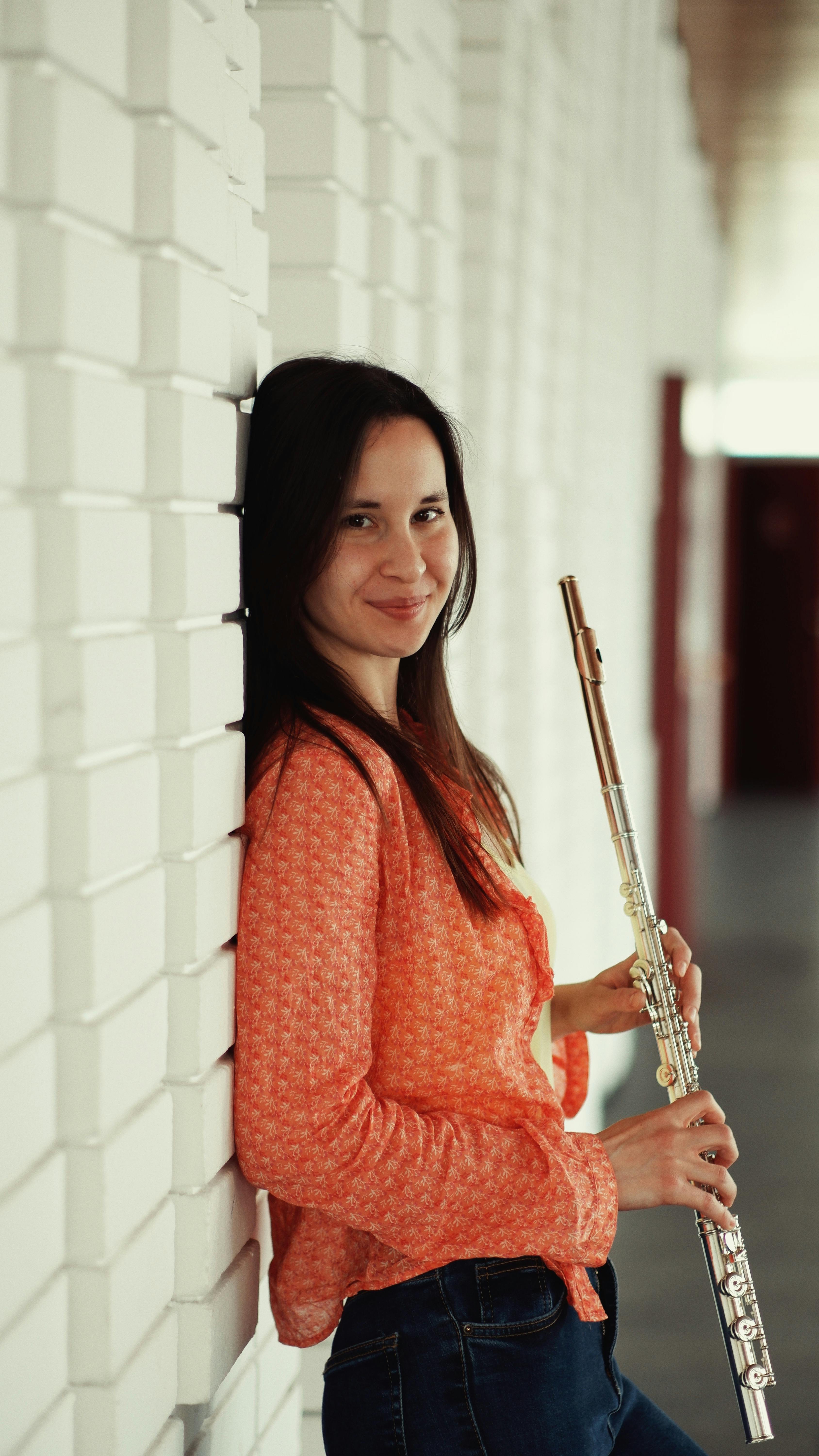 Young woman leaning against white wall, holding a flute, wearing an orange top.