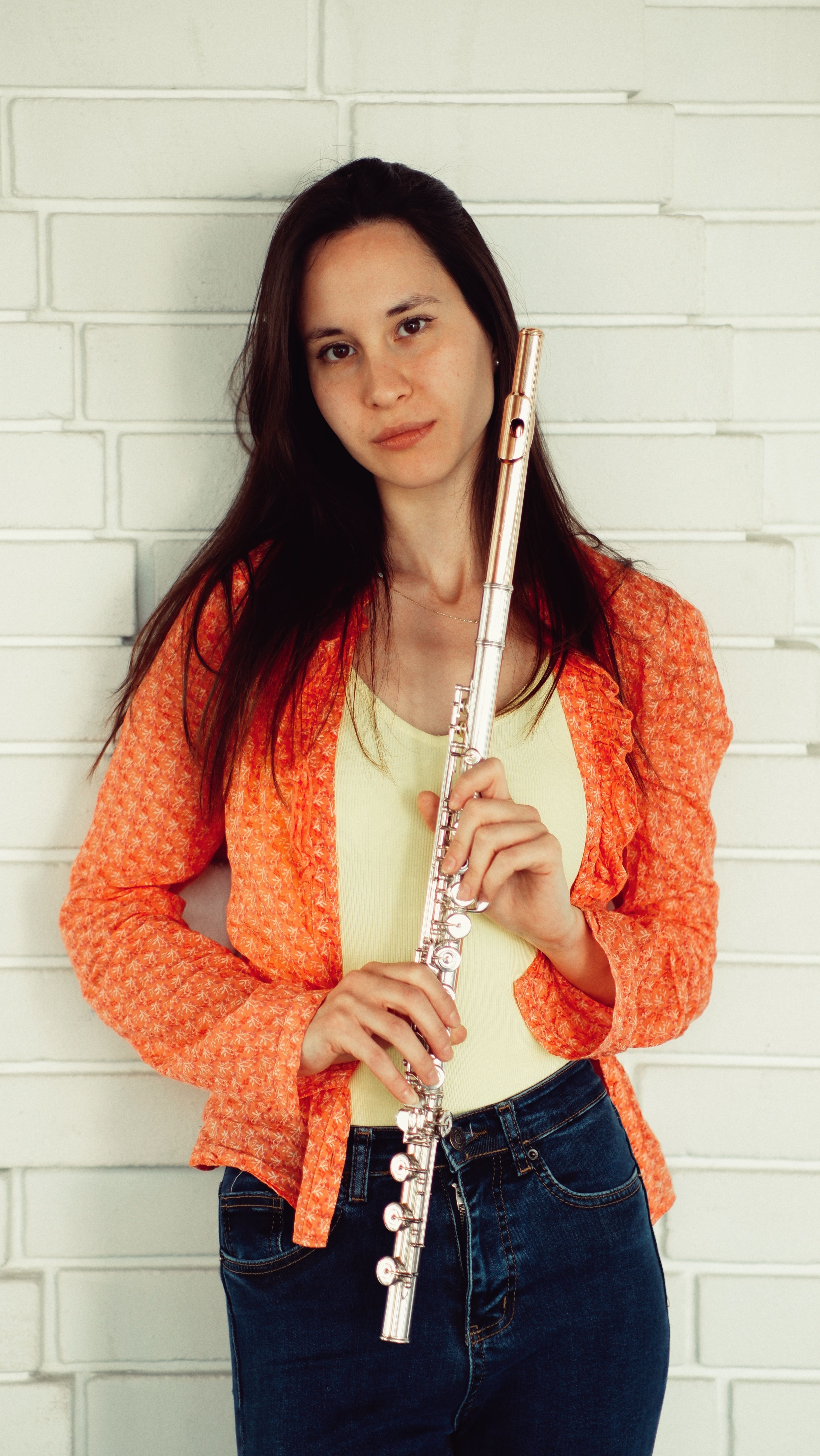A woman in an orange jacket holding a flute poses seriously against a white brick wall.