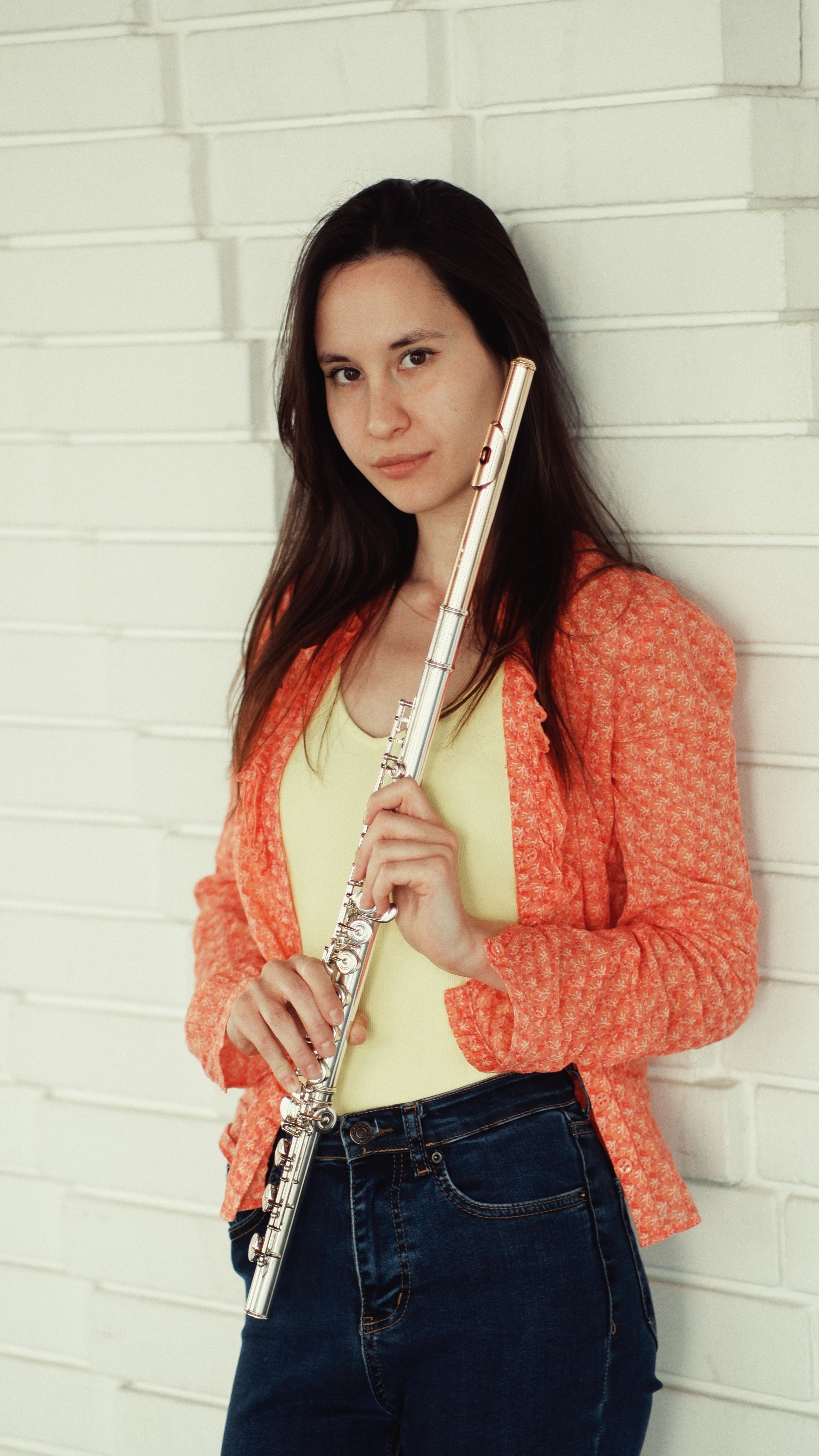 A woman in casual attire holding a flute while leaning on a white brick wall, exuding confidence.