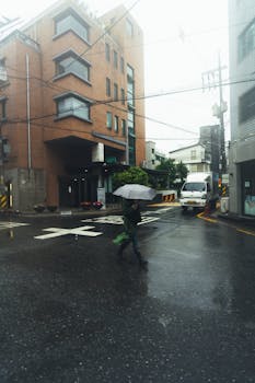 A person walks with an umbrella on a wet city street with modern buildings and a parked van.