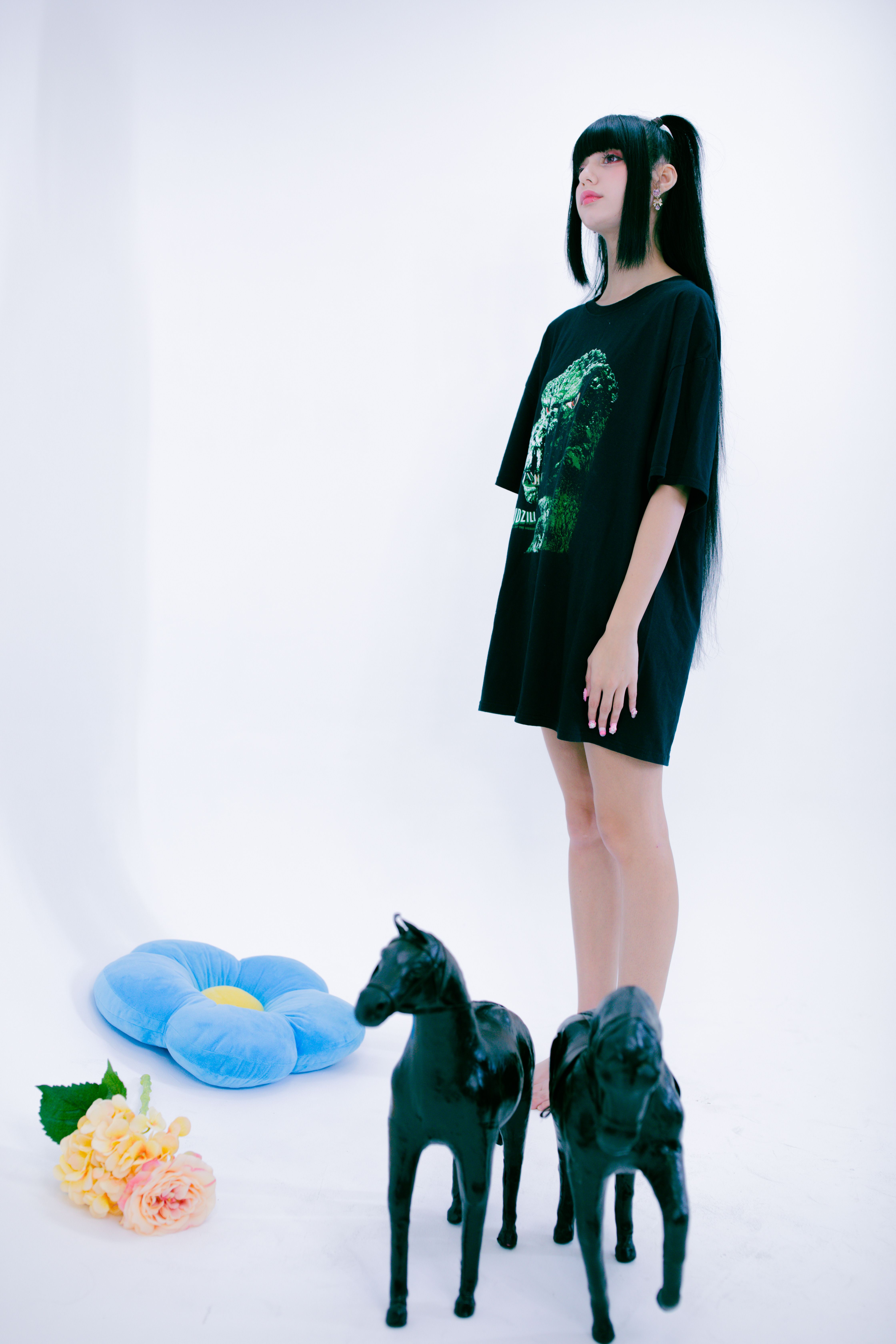 Model in oversized shirt with figurines and flowers in minimalist studio.