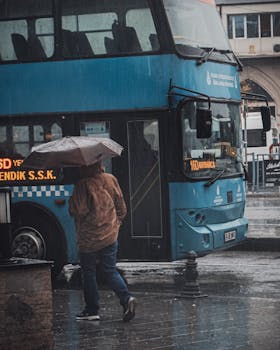 A man with an umbrella walking by a double decker bus on a rainy Istanbul street.