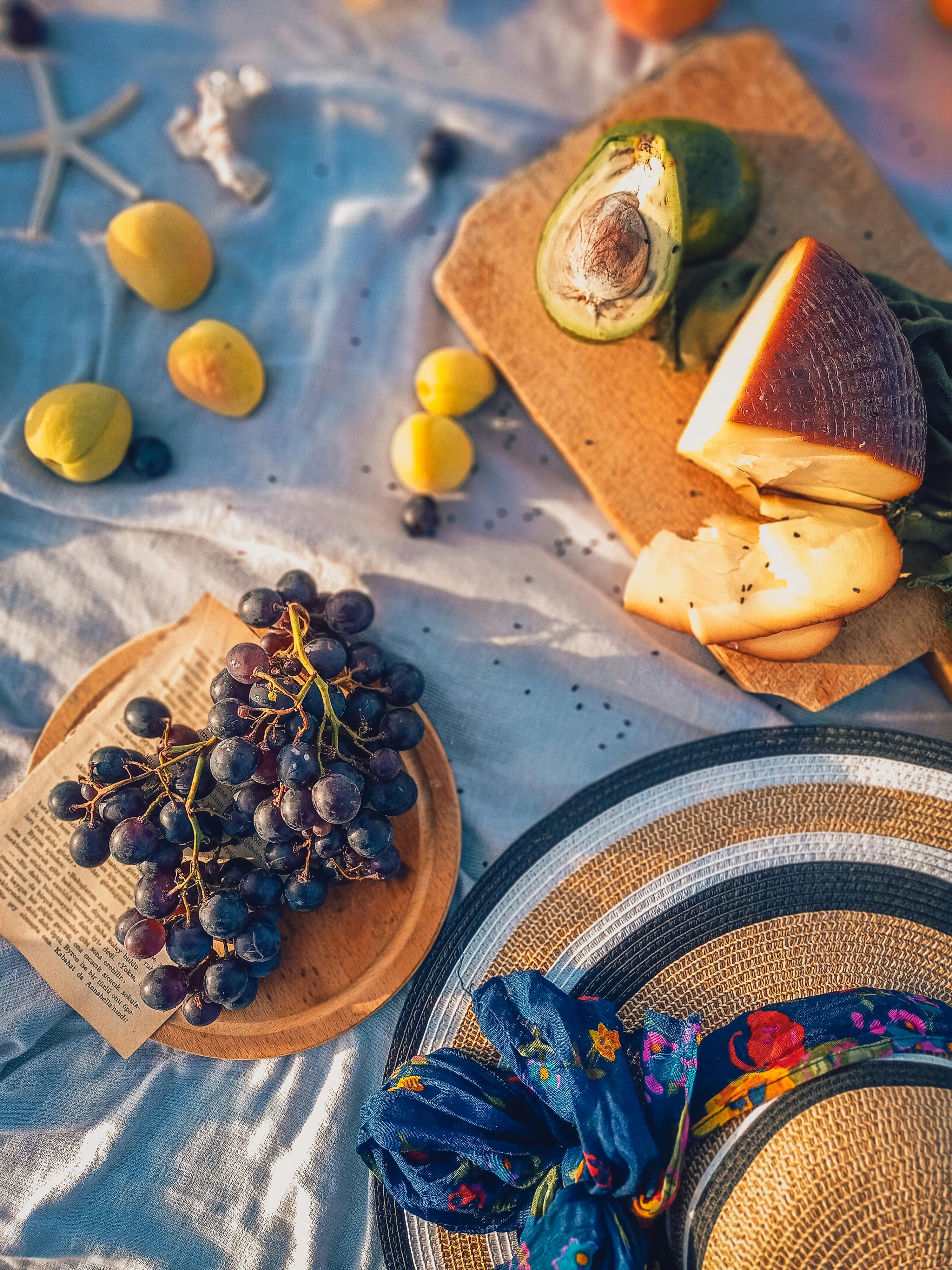 Overhead view of a summer picnic setup with fruits, cheese, and a hat on a cloth.