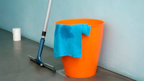 Close-up of cleaning supplies including orange bucket, mop, and blue rag indoors.