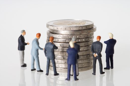 Miniature figurines of businessmen surround a stacked pile of coins on a clean white background.