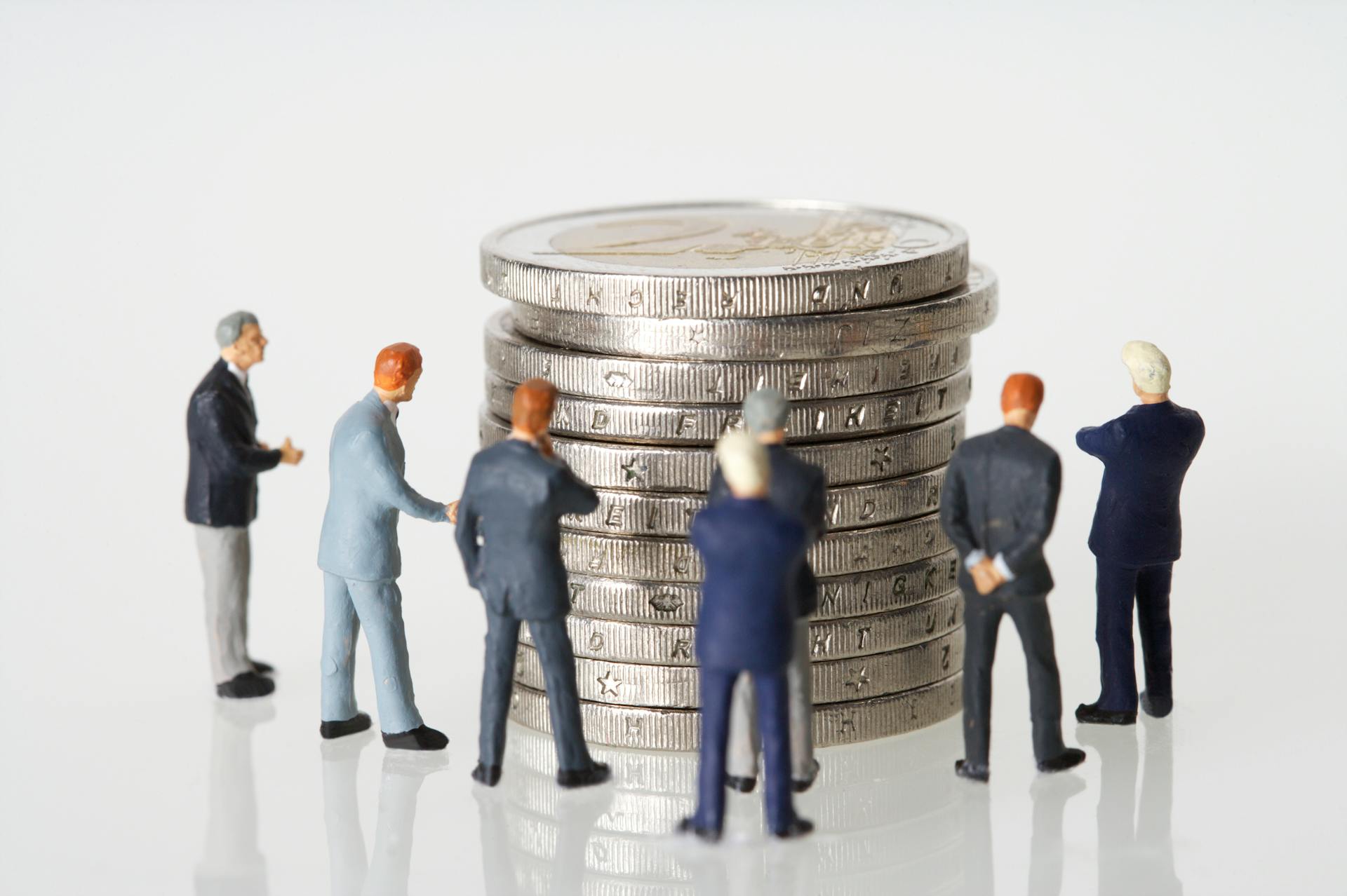 Miniature figurines of businessmen surround a stacked pile of coins on a clean white background.